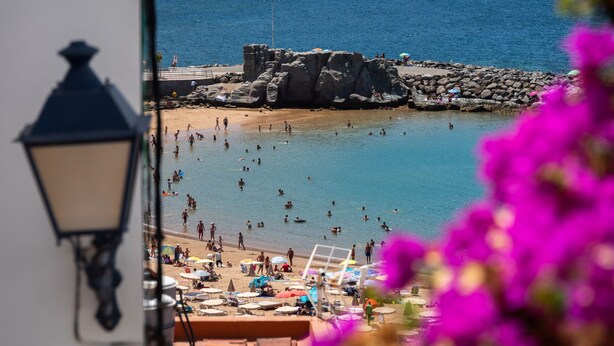 Una spiaggia con persone che nuotano e prendono il sole, una zona rocciosa e un lampione.