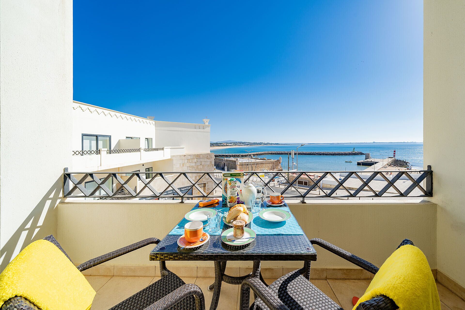 Ein Balkon mit Frühstückstisch, der auf eine Küstenlandschaft mit Leuchtturm und Steg blickt.