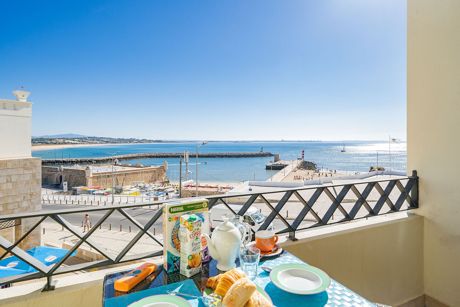 A balcony with a table set for breakfast, overlooking a coastal view with a clear sky.
