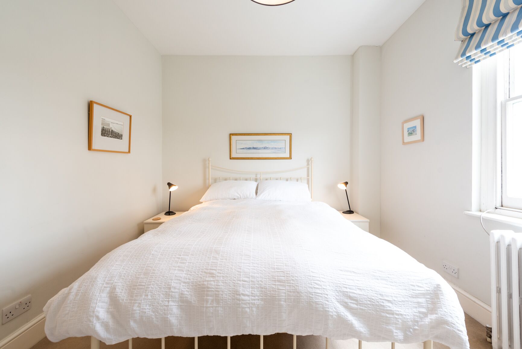 A neatly made bed with white bedding, two bedside tables with lamps, and a window with a striped curtain.