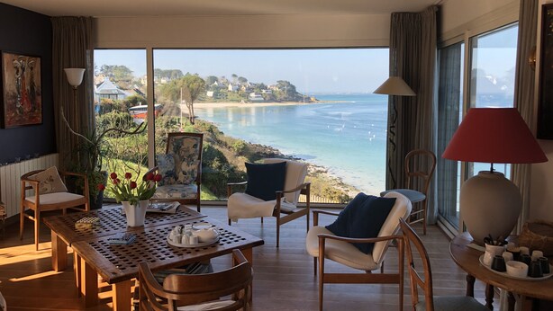 Habitación amplia con un ventanal que da a una playa, muebles de madera y una mesa con un florero.