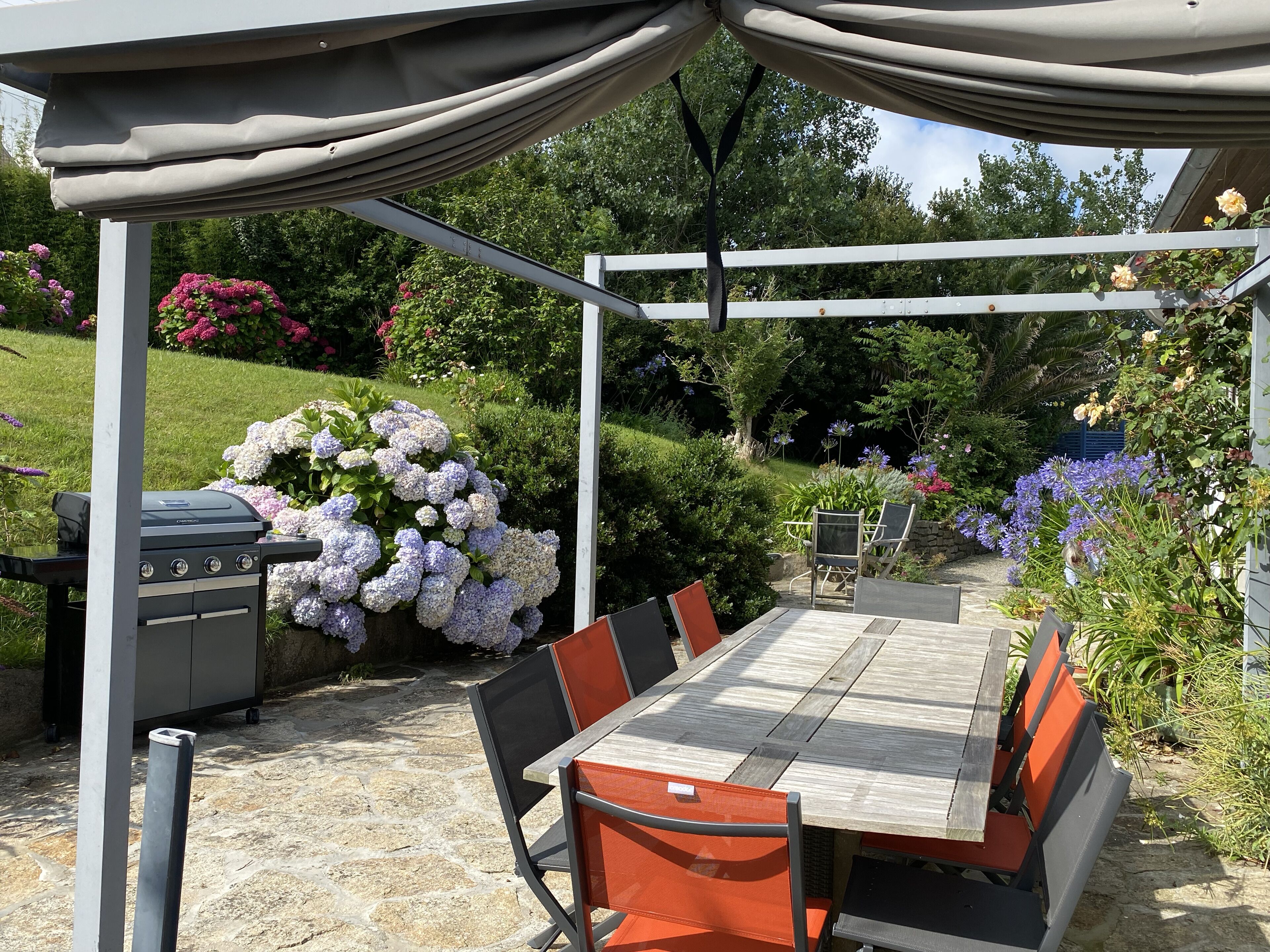 Een terras buiten met een houten tafel en stoelen, een barbecue en een pergola erboven.