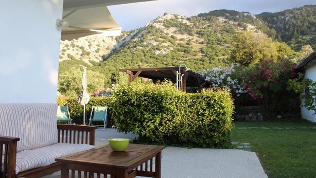 A patio with a wooden table and a cushioned bench, a green bowl, and a view of a mountain landscape.