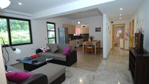 A modern living room with a sofa, coffee table, and a dining area with a table and chairs. A kitchen with appliances and cabinets is visible in the background.