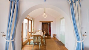 A dining area with a wooden table and chairs, a hanging light fixture, and a view through an arched doorway.