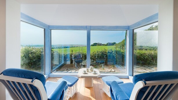 A conservatory with a view of a green field and a distant house. Inside, there is a table with cups and saucers, and two blue cushioned chairs.