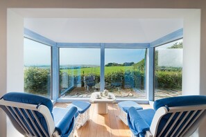 Un invernadero con vista a un campo verde y una casa distante. Adentro, hay una mesa con tazas y platillos, y dos sillones azules acolchados.