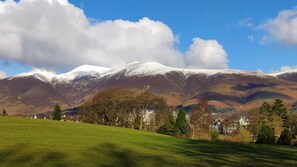 Een bergachtig landschap met besneeuwde toppen, een gazon op de voorgrond en een groepje traditionele huizen.