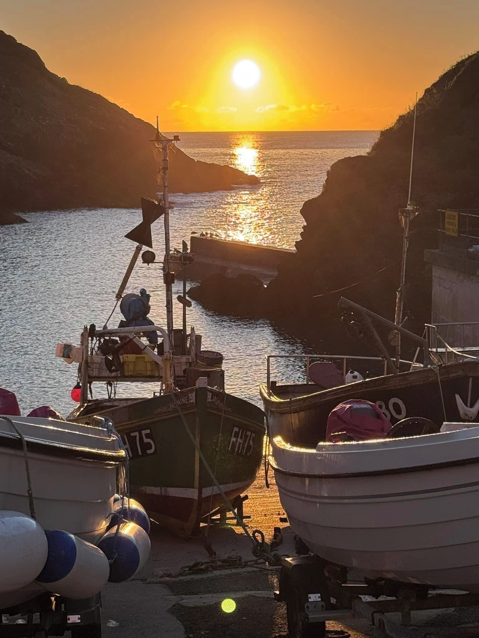 A harbor at sunset with boats docked, a rocky coastline, and a clear sky.