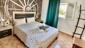Une chambre avec une grande tête de lit en bois en forme de roue, un lit recouvert d’une literie à motifs floraux, une table de chevet avec une lampe et une fenêtre donnant sur un paysage verdoyant.
