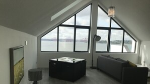 Ein modernes Loft-Zimmer mit schrägem Dach, großen Fenstern mit Blick auf eine Brücke und Wasser, einem dunklen Sofa, einem kleinen Tisch und einer Lampe.