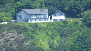 Ein zweistöckiges Haus mit großen Fenstern, gelegen an einem Hang mit Blick auf ein Gewässer.