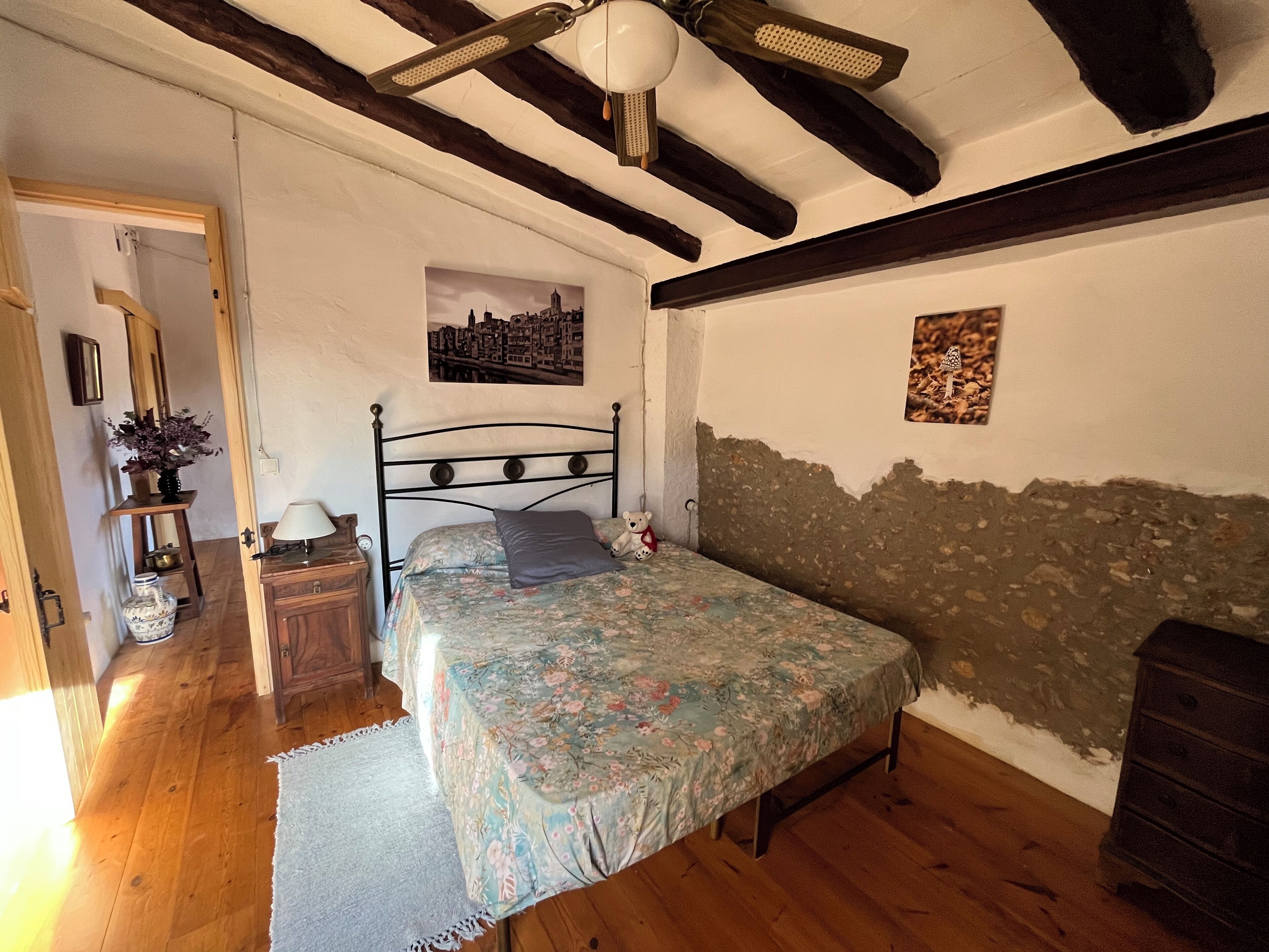 A bedroom with a bed, wooden floor, a ceiling fan, a nightstand, a lamp, a dresser, and a wall with a cityscape painting.