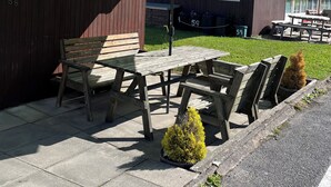 Ein hölzerner Sitzbereich im Freien mit Tisch und Bänken, einem leuchtend gelben Sonnenschirm und einem Gebäude mit Fenstern und einer Tür im Hintergrund.