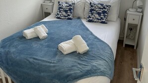 Ein ordentlich bezogenes Bett mit blauer Decke und Kissen, eine Nachttischlampe und ein Fenster mit durchsichtigem Vorhang.