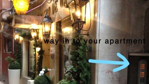 A street view with a building entrance, potted plants, and a sign indicating the way to an apartment.