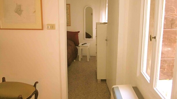 A narrow hallway leading to a bedroom with a bed, a chair, and a desk. A framed picture is on the wall, and there is a window on the right side.
