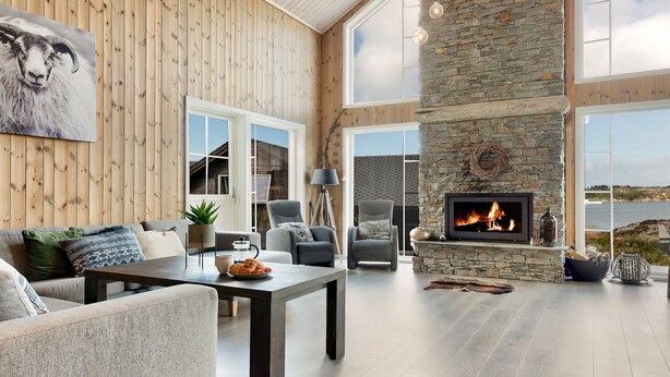 A spacious living room with a stone fireplace, wooden ceiling beams, and large windows offering a view of the outdoors.