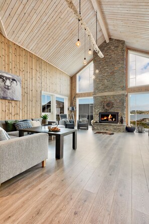A spacious living room with a stone fireplace, wooden ceiling beams, and large windows offering a view of the outdoors.