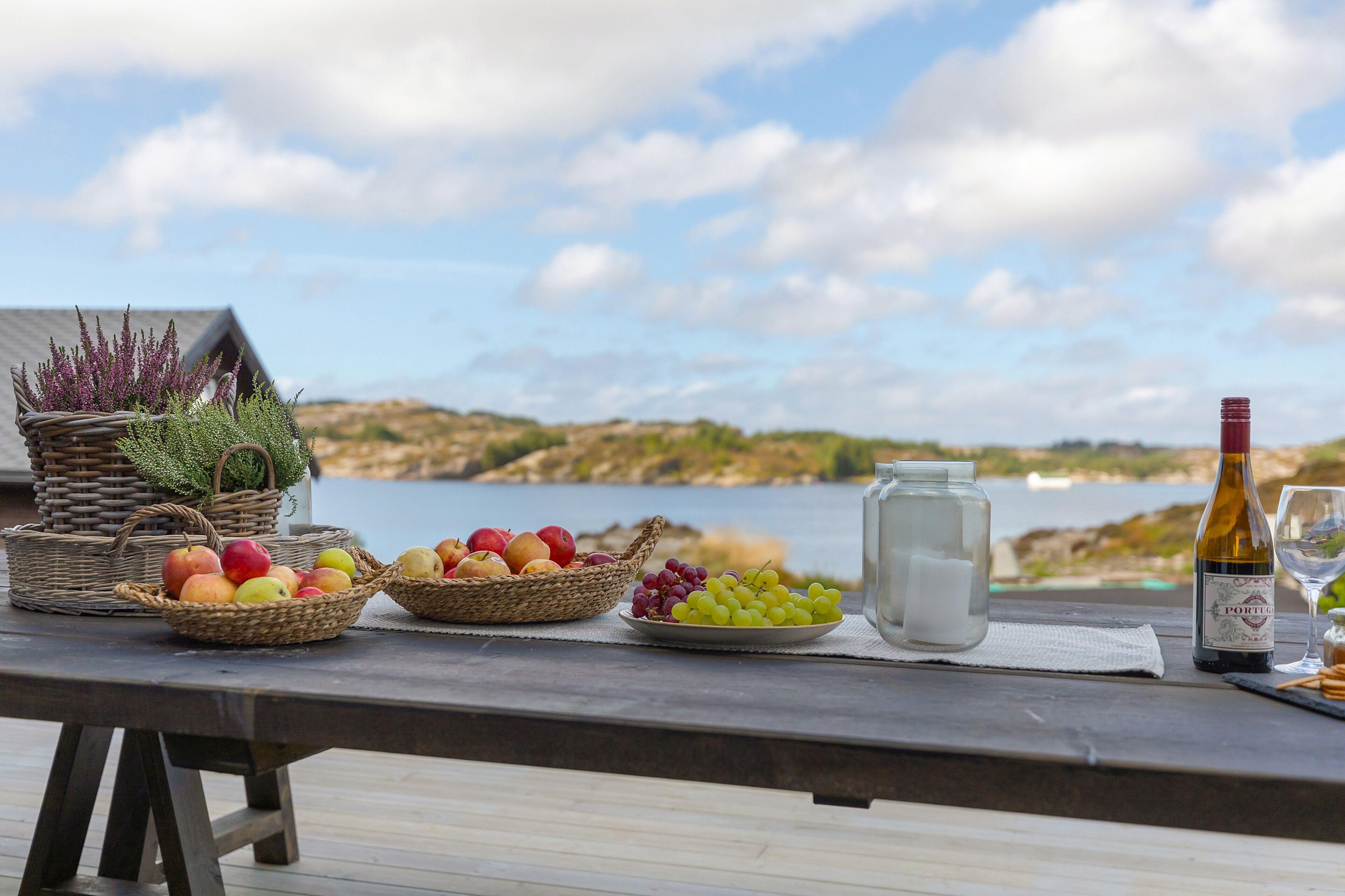Une table en bois, sur laquelle on a disposé des paniers de fruits, une bouteille de vin et un verre, se trouve sur une terrasse donnant sur un plan d’eau.