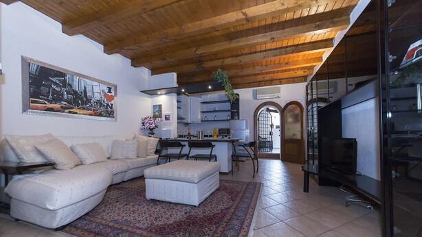 A modern living room with a sectional sofa, a coffee table, and a rug. There is a kitchen area with a dining table and chairs, and a TV on a stand.