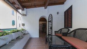 A covered outdoor seating area with a glass table and black wicker chairs, a white wall with a window, and a tiled floor.