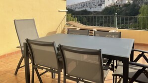 En balkon med et bord og stole, der giver udsigt over et boligområde med hvide bygninger og grønt.