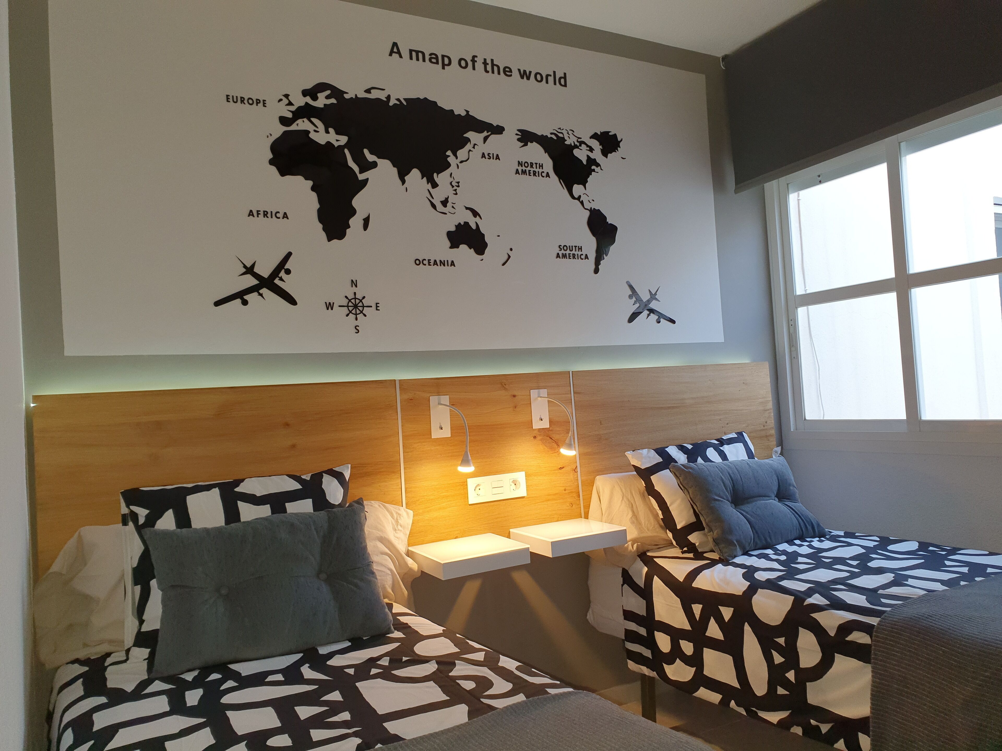 Habitación de hotel con un mapa del mundo en la pared, dos camas con ropa de cama estampada, mesitas de noche con lámparas y una ventana con luz natural.