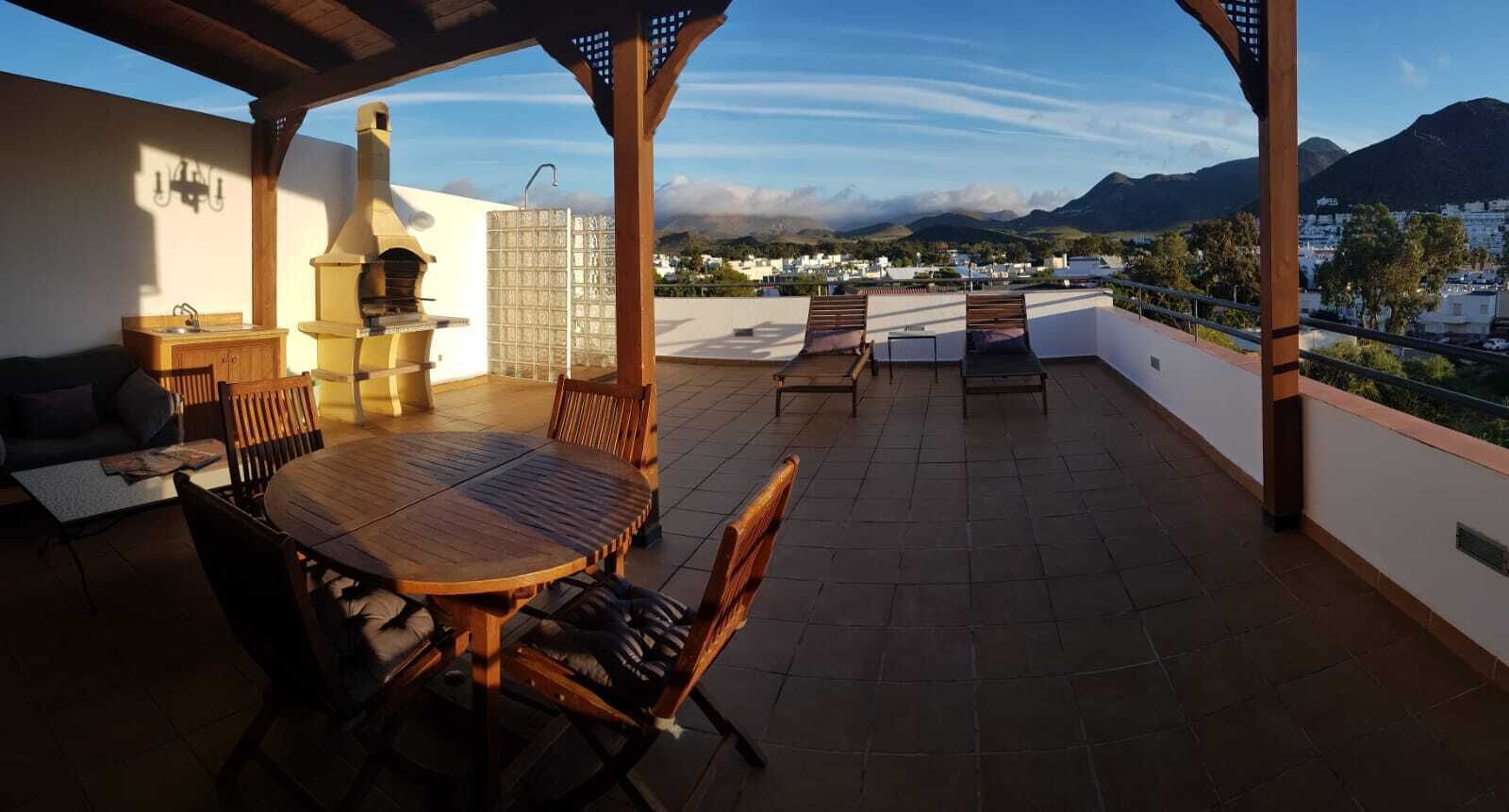 A rooftop terrace with a wooden table and chairs, a grill, and a view of the city and mountains.