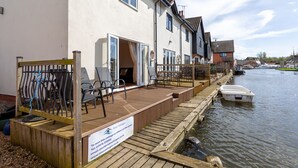 A riverside property with a wooden deck, outdoor seating, and a sign indicating rental availability.