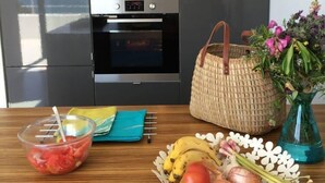 Una cocina con electrodomésticos modernos, una mesa de madera con frutas y verduras frescas, una cesta tejida y un jarrón con flores.