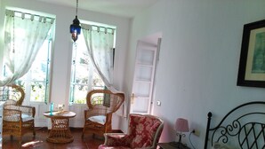 Una habitación con una cama, dos sillas de mimbre, una mesita, un sillón rojo con estampado floral, una lámpara y una puerta.