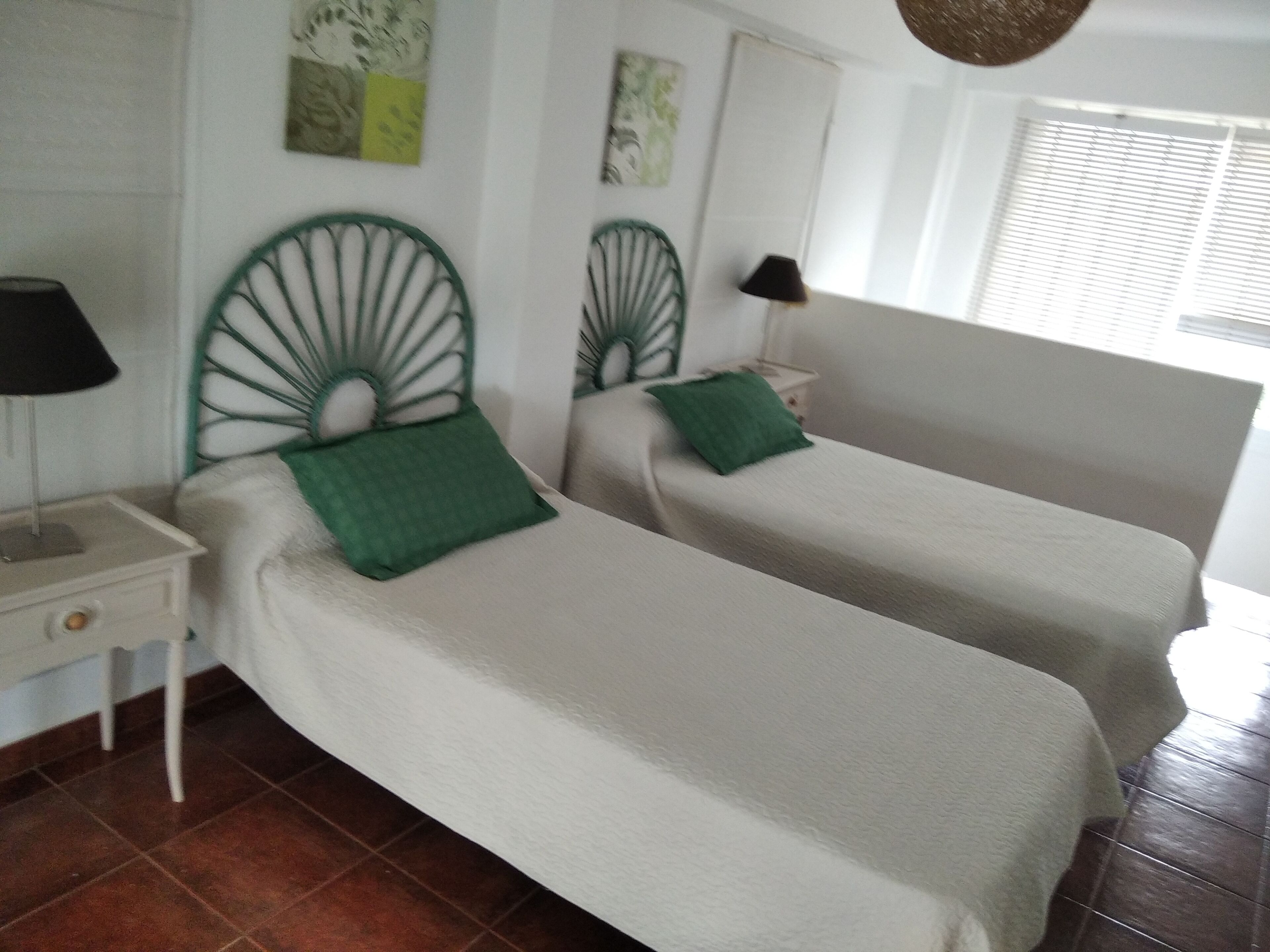A room with two single beds, each with a green pillow and a white headboard. There is a nightstand with a lamp on the left side. The room has a tiled floor and a window with blinds.