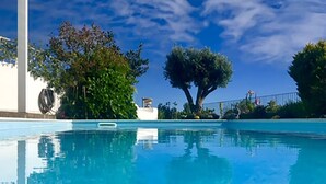Une piscine d’un bleu éclatant, avec une structure blanche sur la gauche et des arbres en arrière-plan.