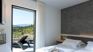 A modern hotel room with a balcony, a bed with white linens, and a view of a forested area. - Villa Blanche - Top quality villa surrounded by nature, close to the beach (Chania)