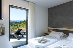 A modern hotel room with a balcony, a bed with white linens, and a view of a forested area.
