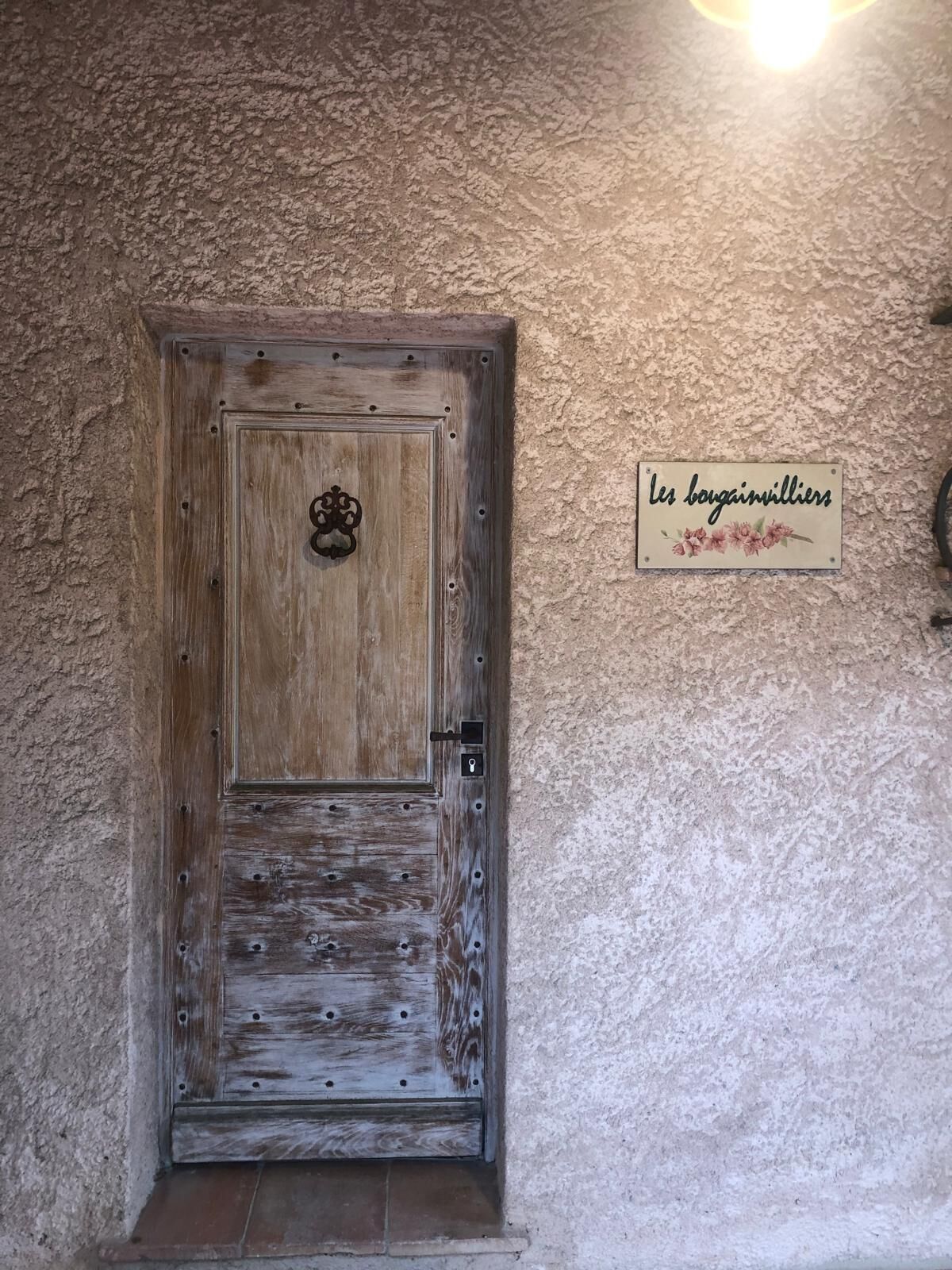 Eine rustikale Holztür mit einem Metallgriff und Schlüsselloch, in eine strukturierte Wand eingelassen, mit einem Schild, auf dem 'les bougainvilleis' steht.