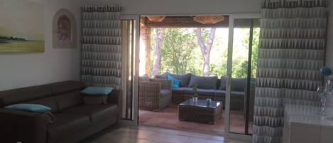 Un salon avec un canapé marron, une table basse en osier et une porte en verre donnant sur un espace extérieur avec vue sur les arbres.