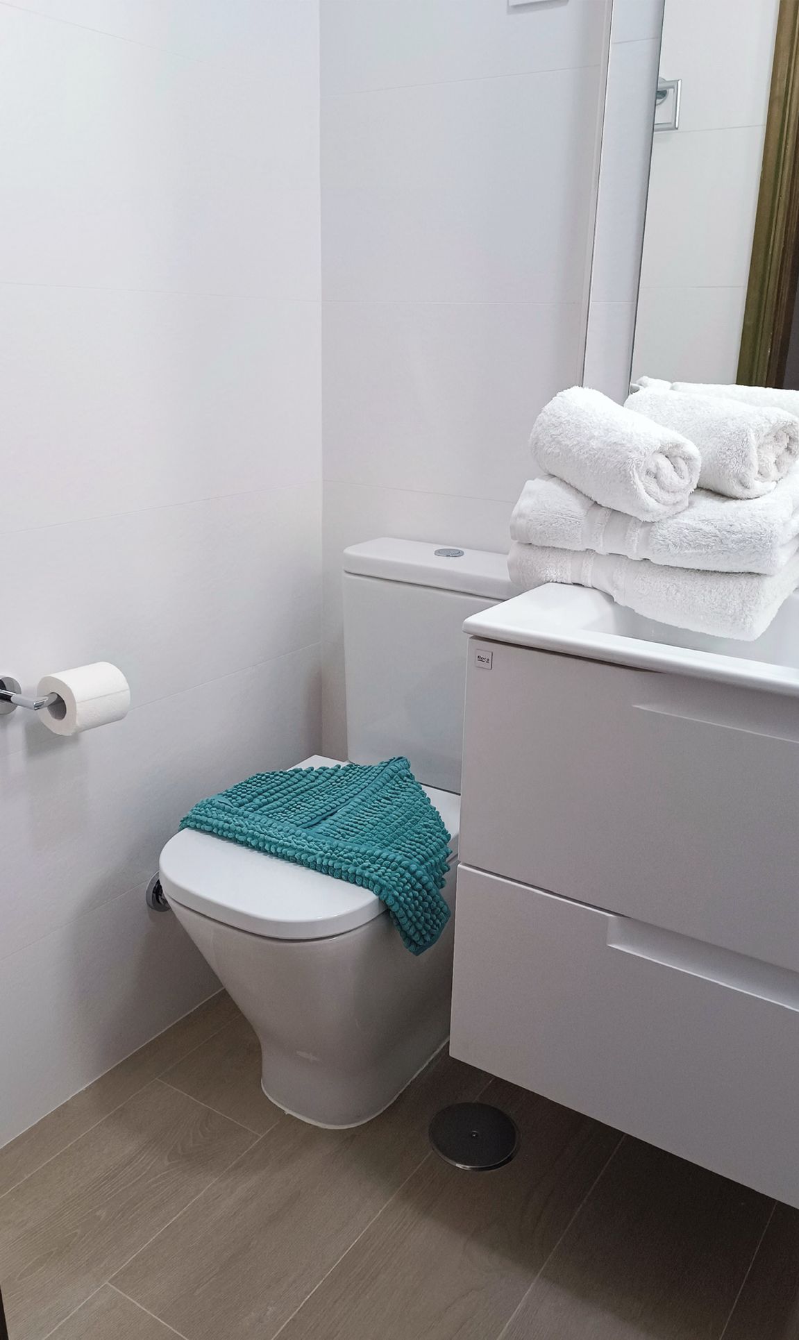 Un baño pequeño y limpio con inodoro, una pila de toallas blancas, una alfombrilla de baño color turquesa y un rollo de papel higiénico.
