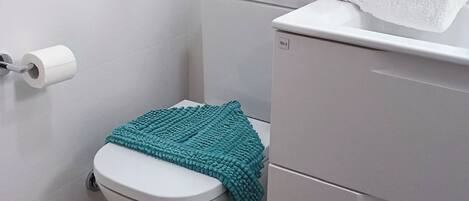 Un baño pequeño y limpio con inodoro, una pila de toallas blancas, una alfombrilla de baño color turquesa y un rollo de papel higiénico.