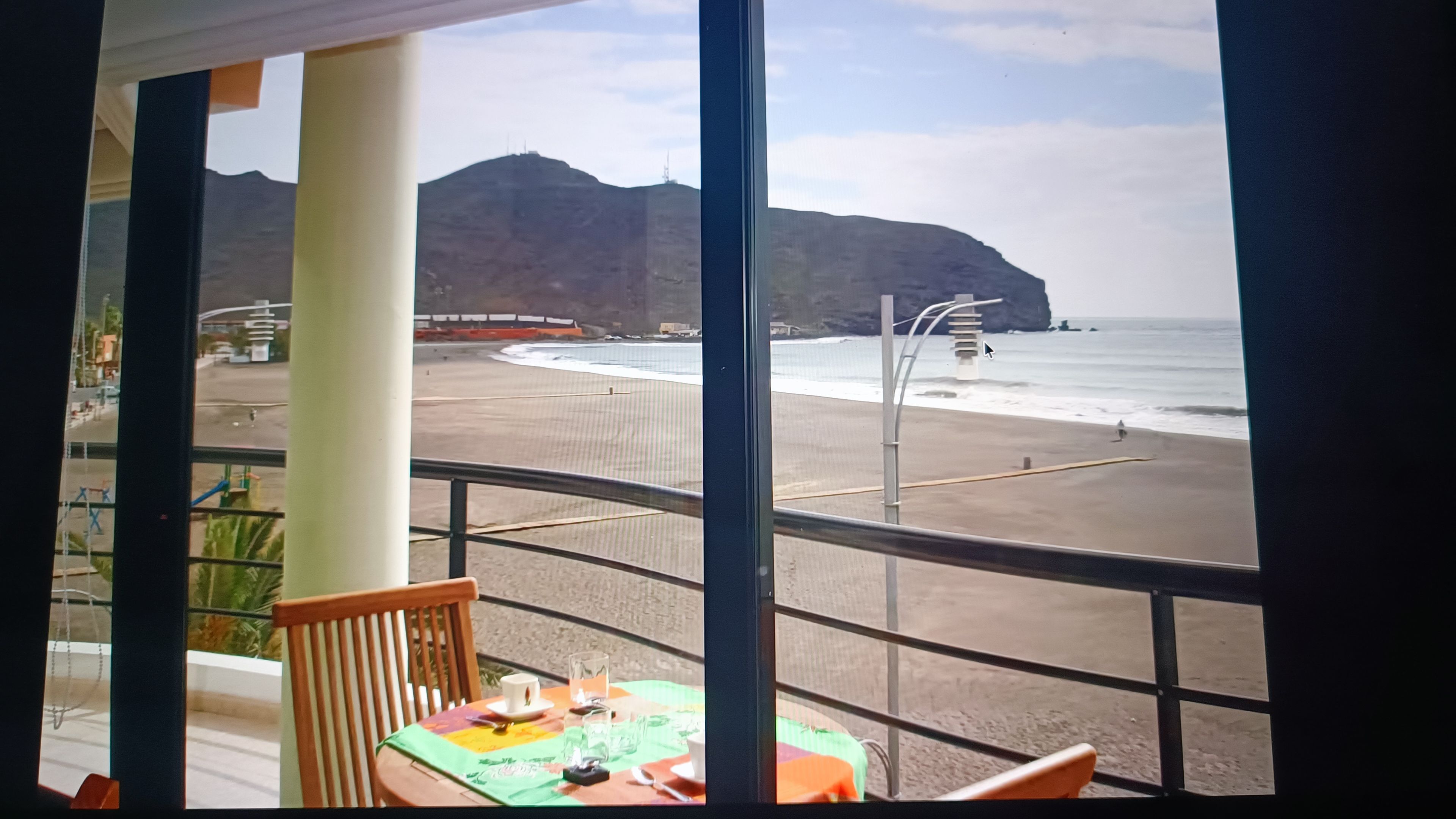 A table with a tablecloth, a chair, and a view of a beach with mountains in the background.