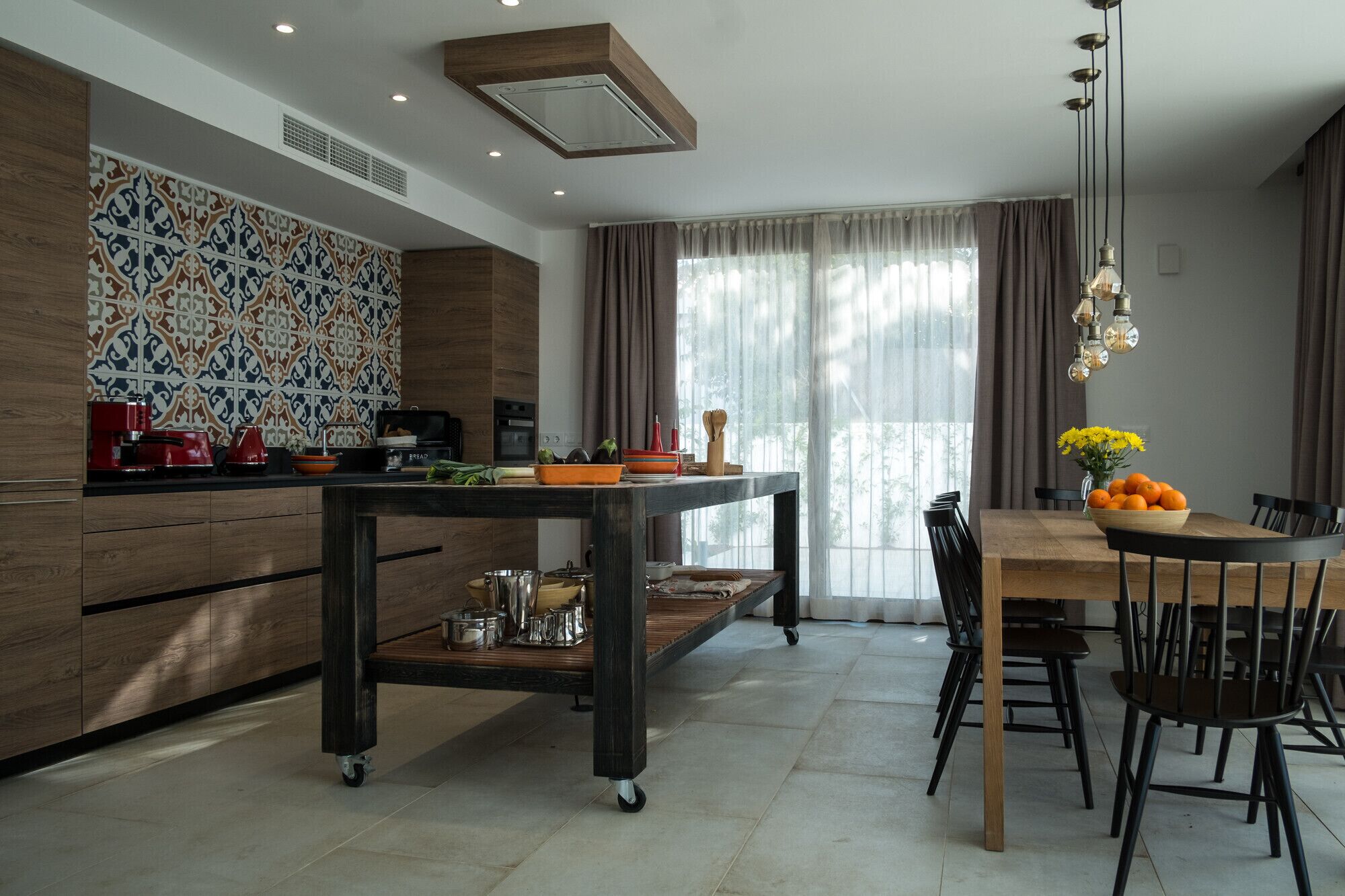 A modern kitchen with a dining area, featuring a kitchen island, wooden cabinets, and a large window with curtains.