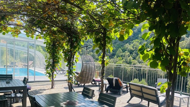An outdoor patio with wooden tables and chairs, a hanging chair, and a view of a pool and greenery.