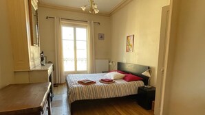 Een slaapkamer met een bed, een dressoir, een stoel, een nachtkastje en een raam met gordijnen.
