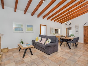 Un salón amplio con un sofá gris, vigas de madera, suelo de baldosas y un comedor con mesa y sillas de madera.
