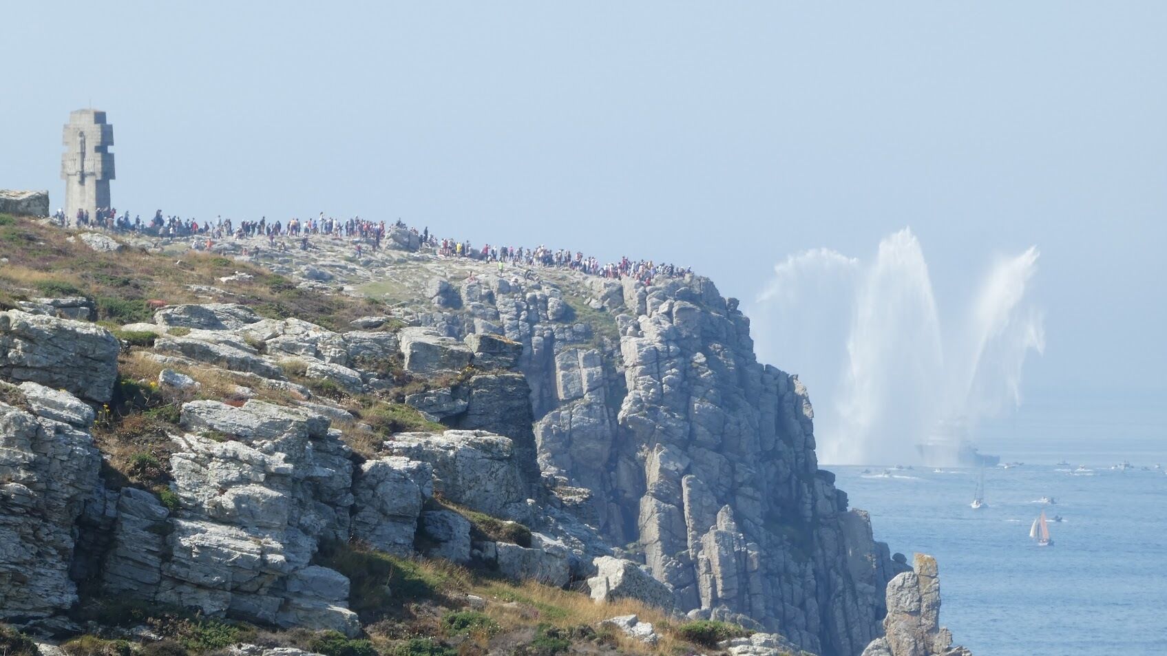 A rocky coastal landscape with a stone tower and a crowd of people on a cliff overlooking the sea.