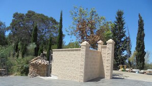 Un mur de pierre surmonté d’une toiture en tuiles, entouré d’arbres et bordé d’une zone pavée.