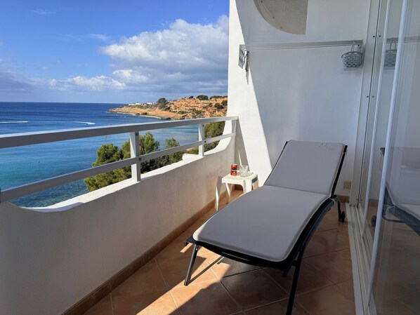 A balcony with a white chair, a small table with a drink, and a view of the sea and buildings.