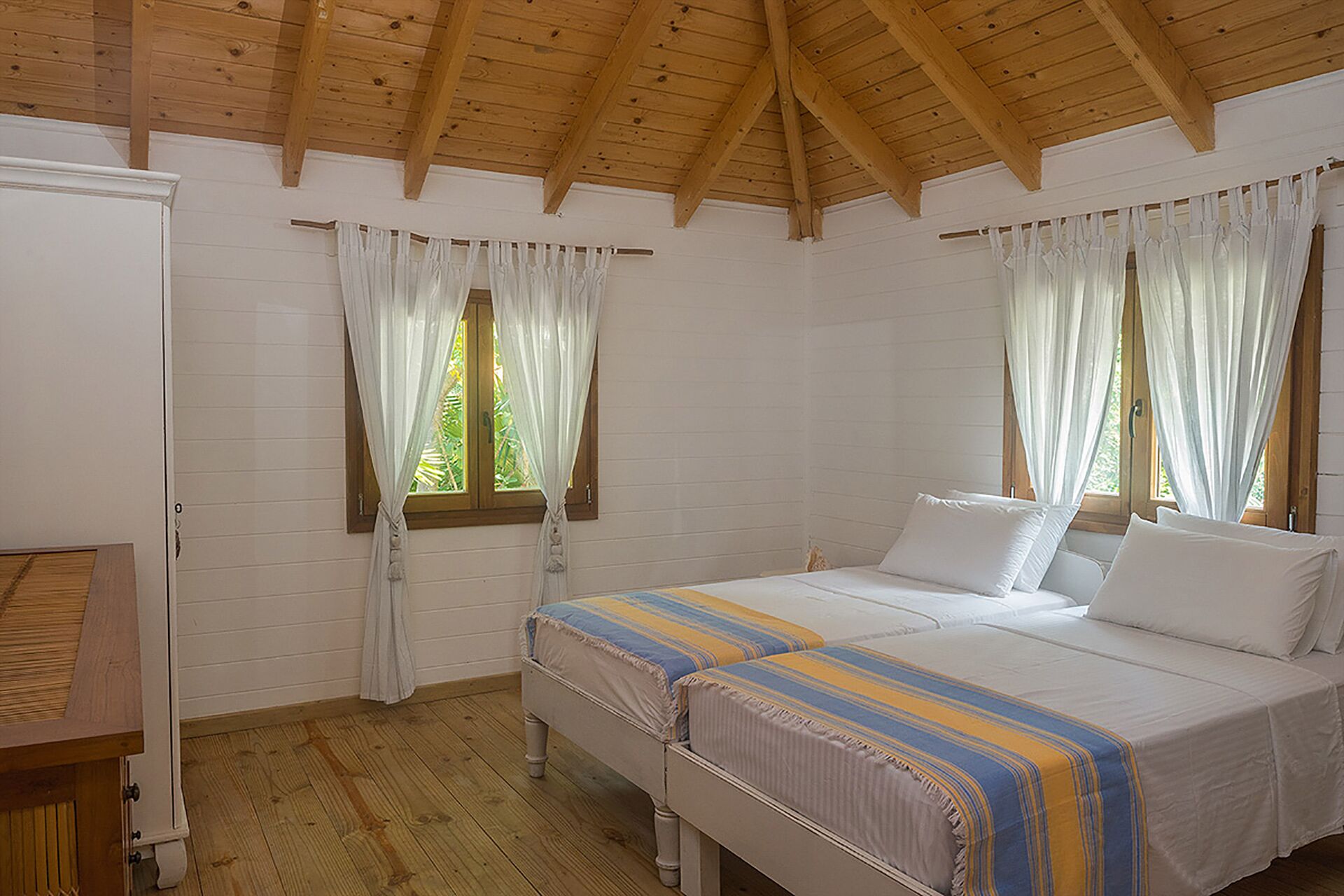 A bedroom with two beds, wooden ceiling beams, white walls, and windows with white curtains.