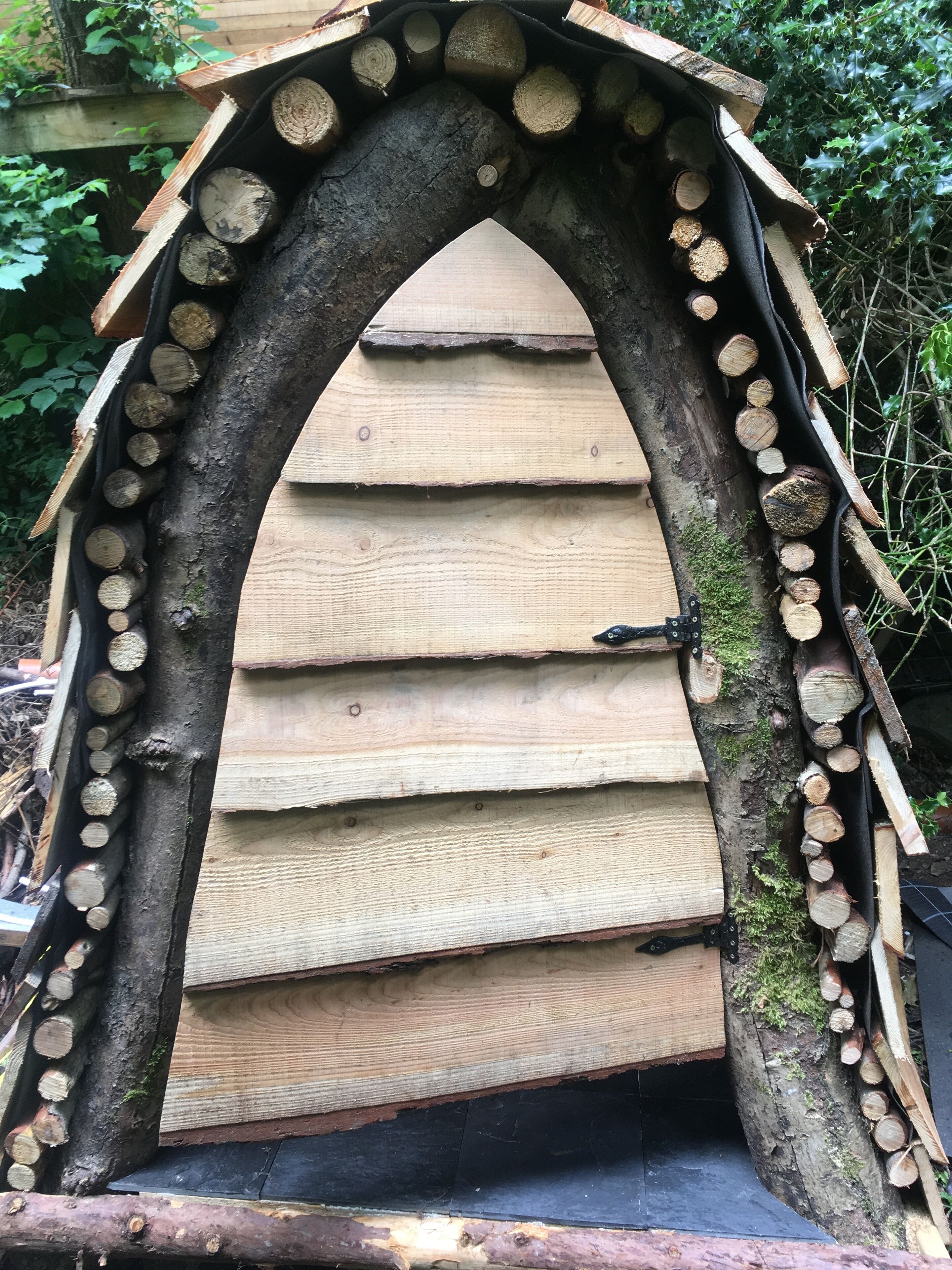 Una estructura de madera con techo inclinado y una puerta, rodeada de troncos apilados.